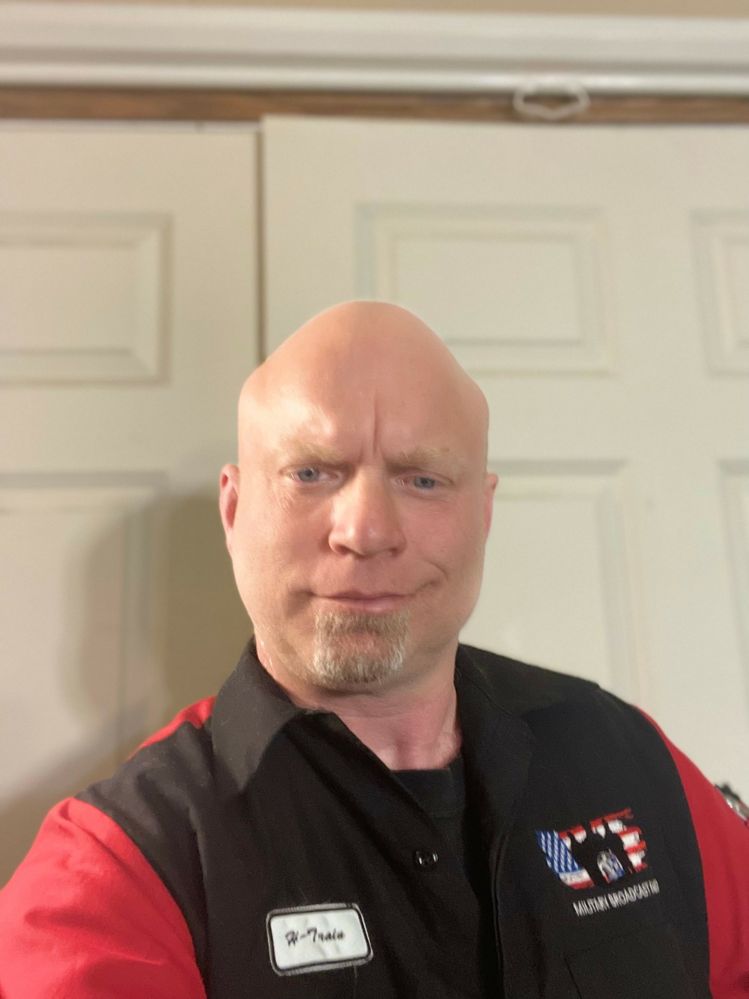 Man with a serious expression, bald head, wearing a black and red uniform with a name tag, standing in front of white doors.