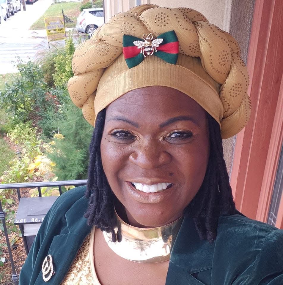 Woman smiling outdoors wearing a beige headwrap with a bow and a green jacket.