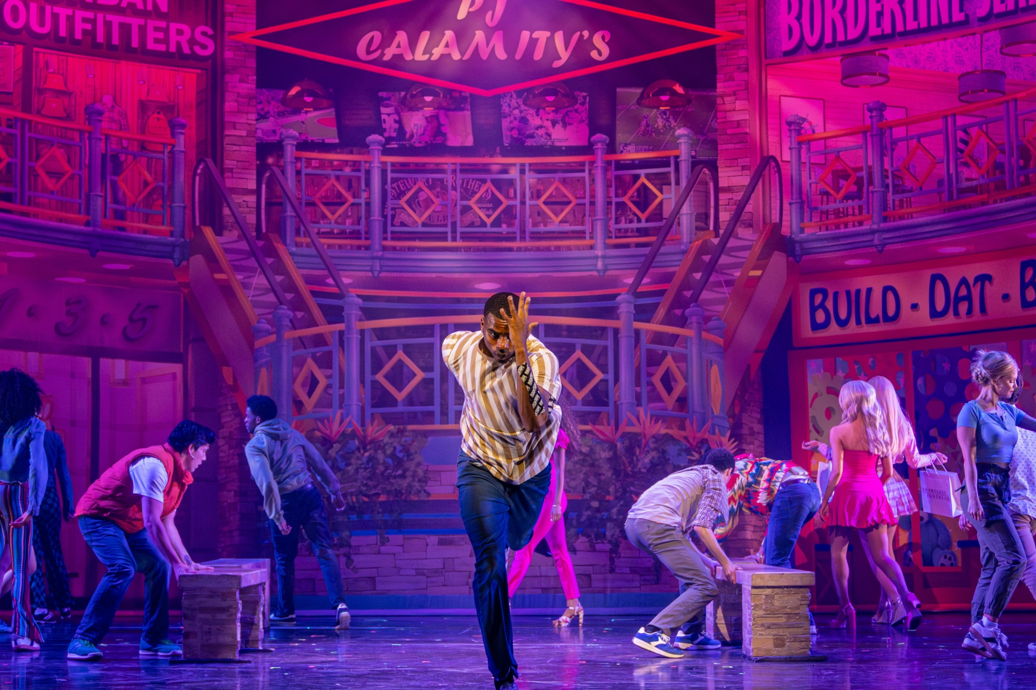 Group of people on stage with colorful set, some bending over, others standing, in a theatrical performance scene.