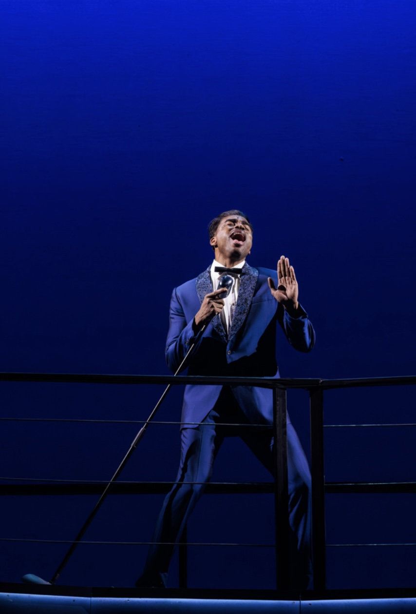 Man in a suit singing on stage with a blue background, holding a microphone, standing behind a railing.