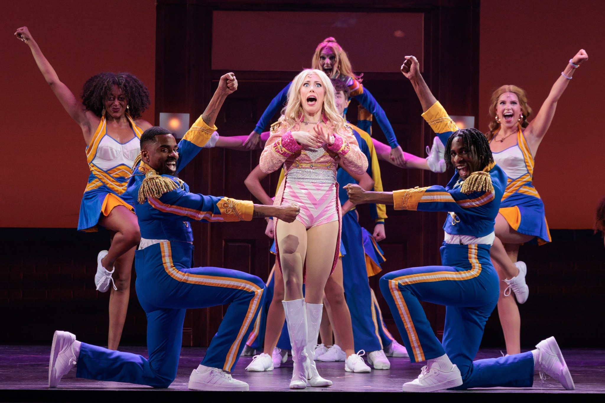 Group of performers on stage, some kneeling and others standing, with arms raised and expressive poses, colorful costumes, and a red background.