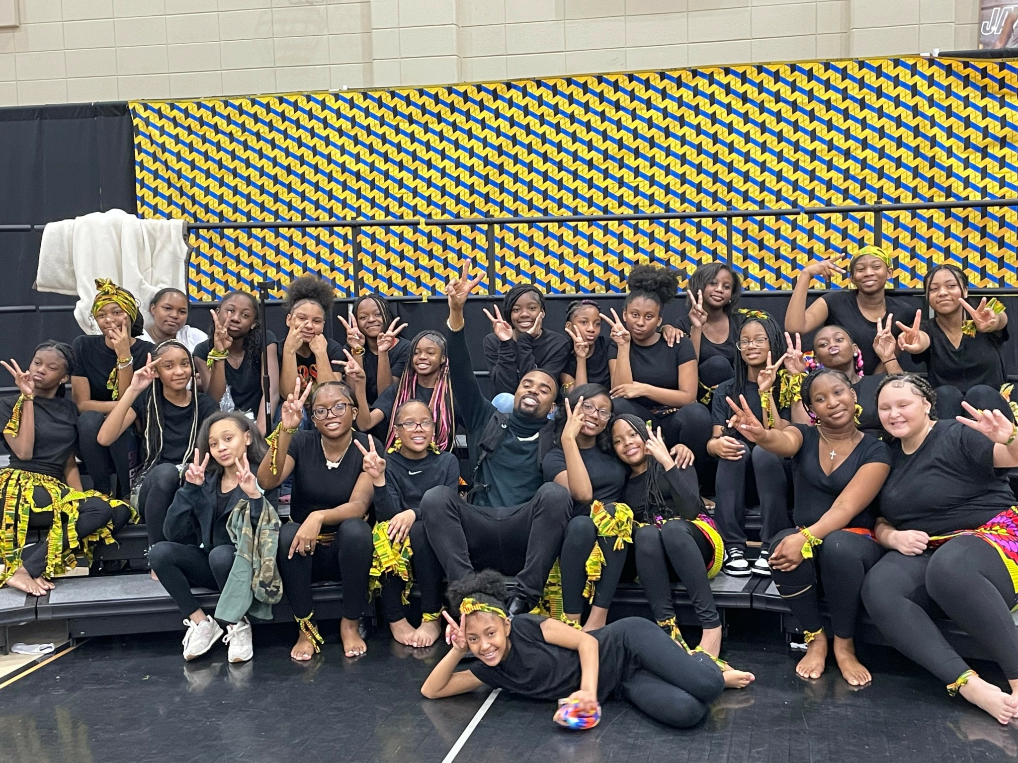 Group of people in black outfits with yellow accessories, posing on stage with hands raised, smiling, in front of a patterned backdrop.