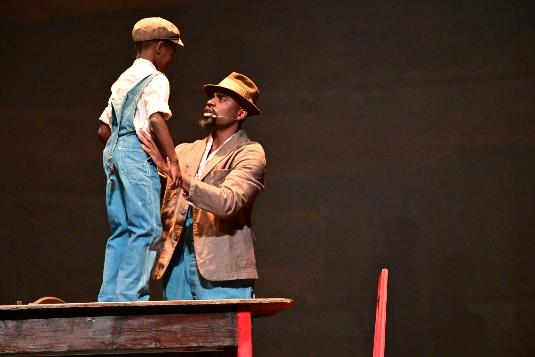 Man kneeling and holding a child's hands on stage, both wearing hats, with a dark background.