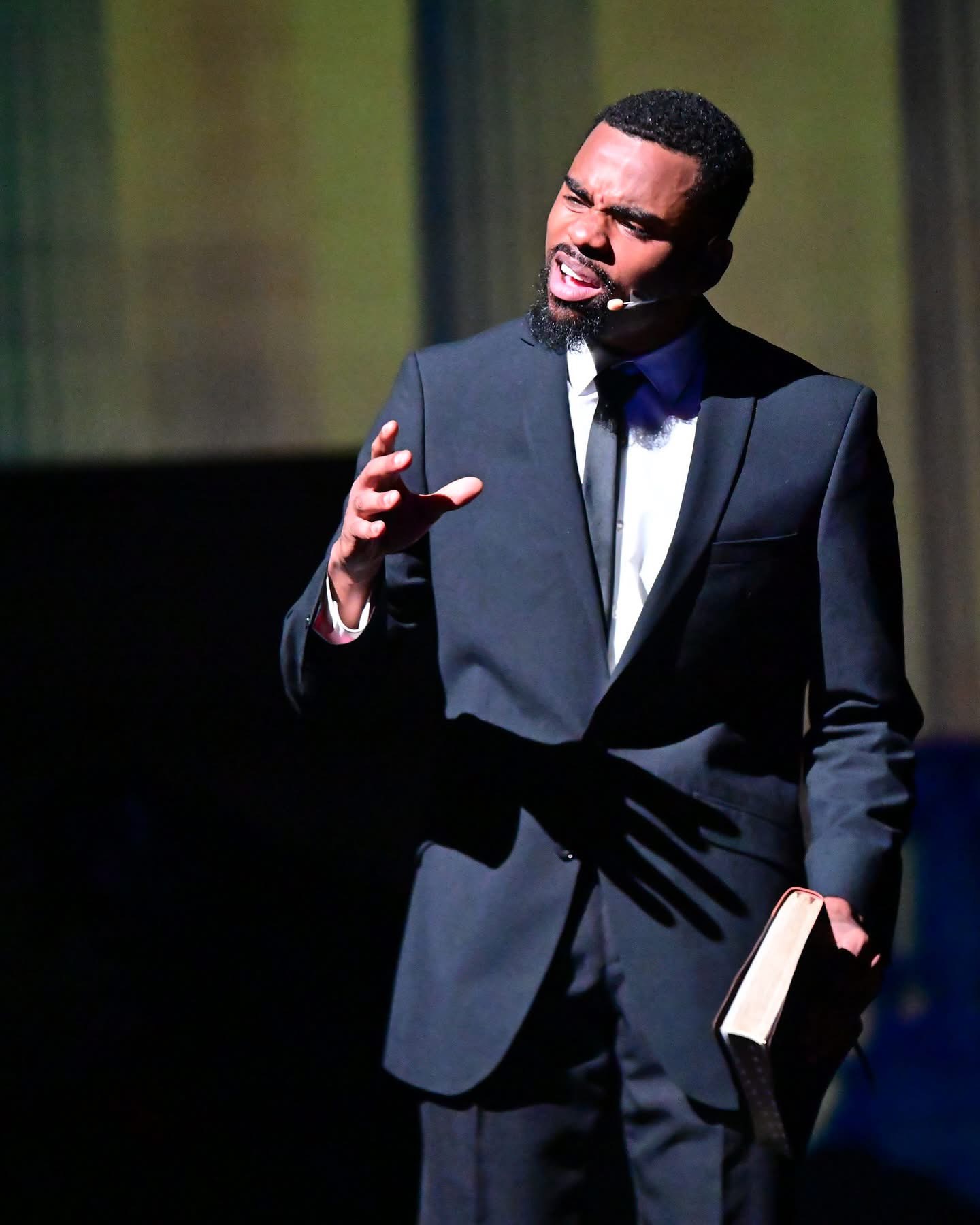 Man in a suit speaking on stage, holding a book, with a dark background and curtain, gesturing with his hand.