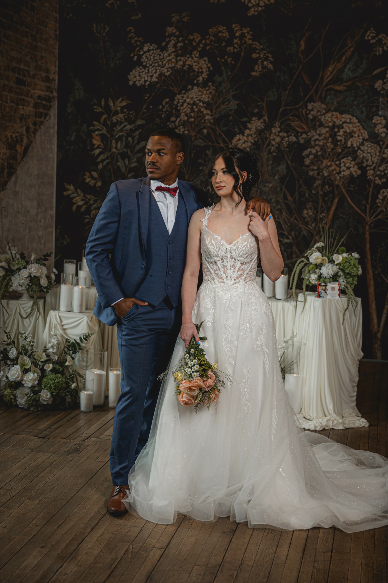 A bride and groom stand together indoors, with floral arrangements and candles in the background, dressed in wedding attire.