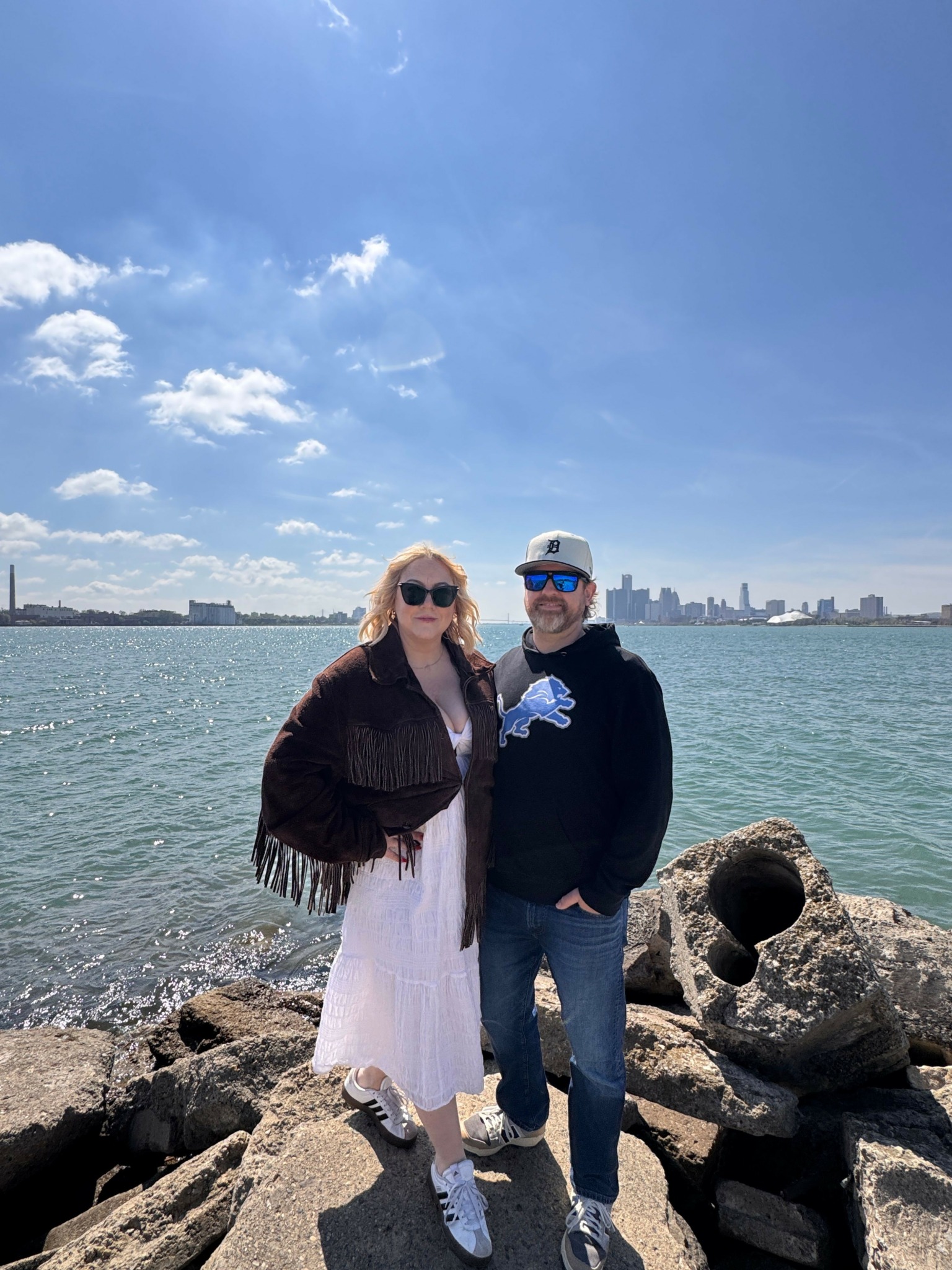 Two people stand on rocks by water with a city skyline in the distance under a partly cloudy sky.