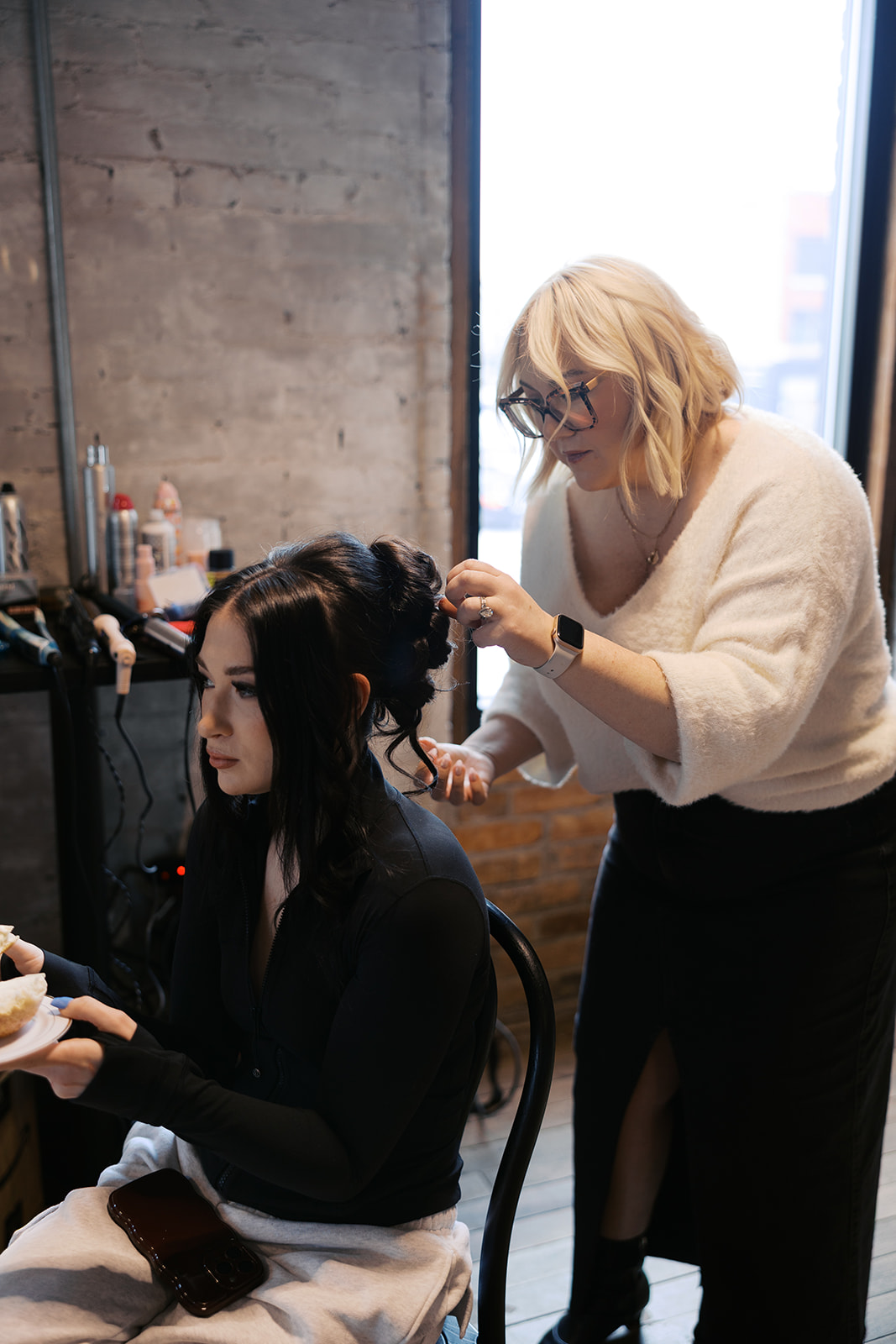 Hairdresser styles a woman's hair in a salon with a concrete wall and large window behind them.