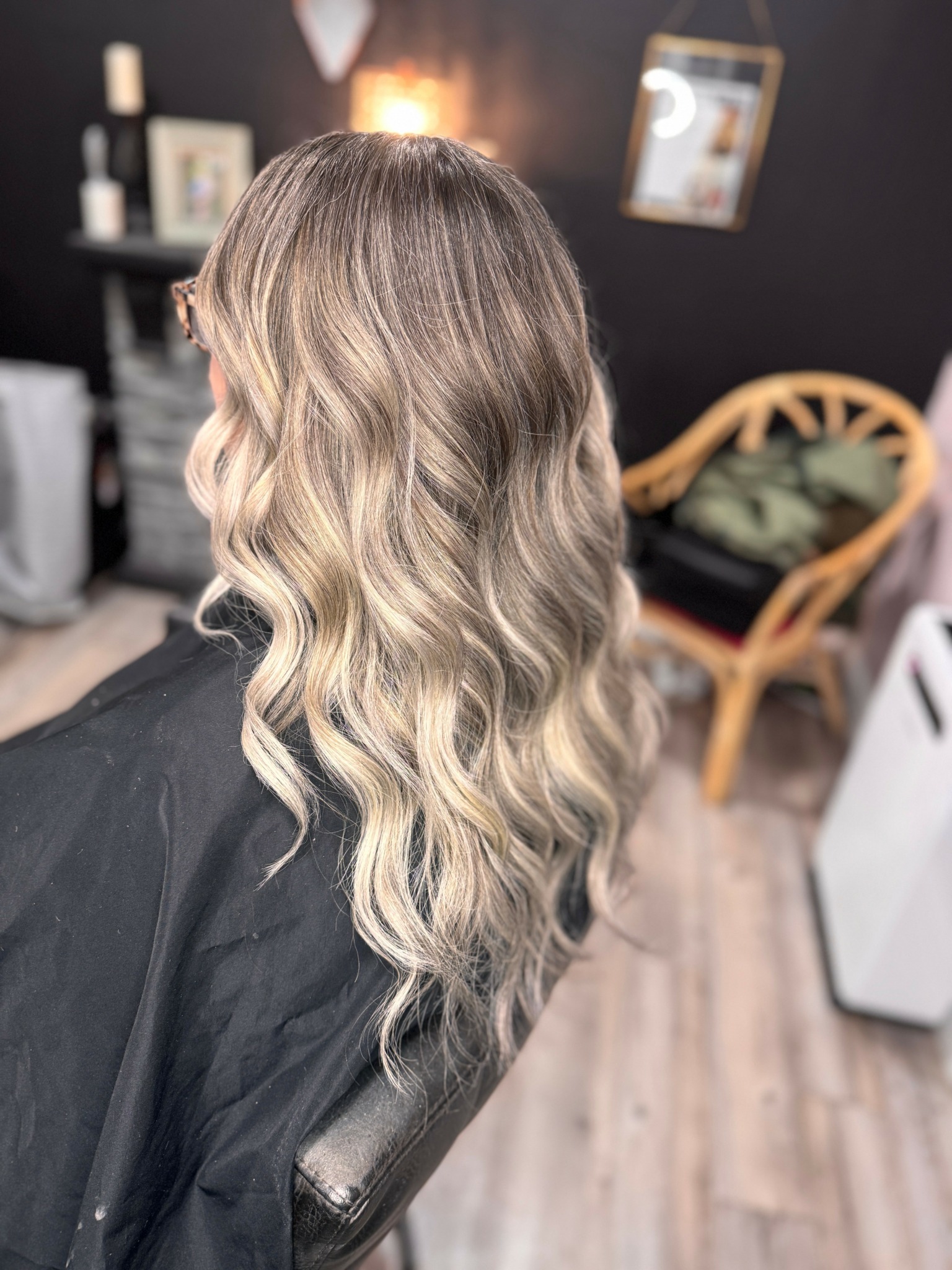 Back view of a woman with long, wavy, blonde hair in a salon, with a chair and mirror in the background.
