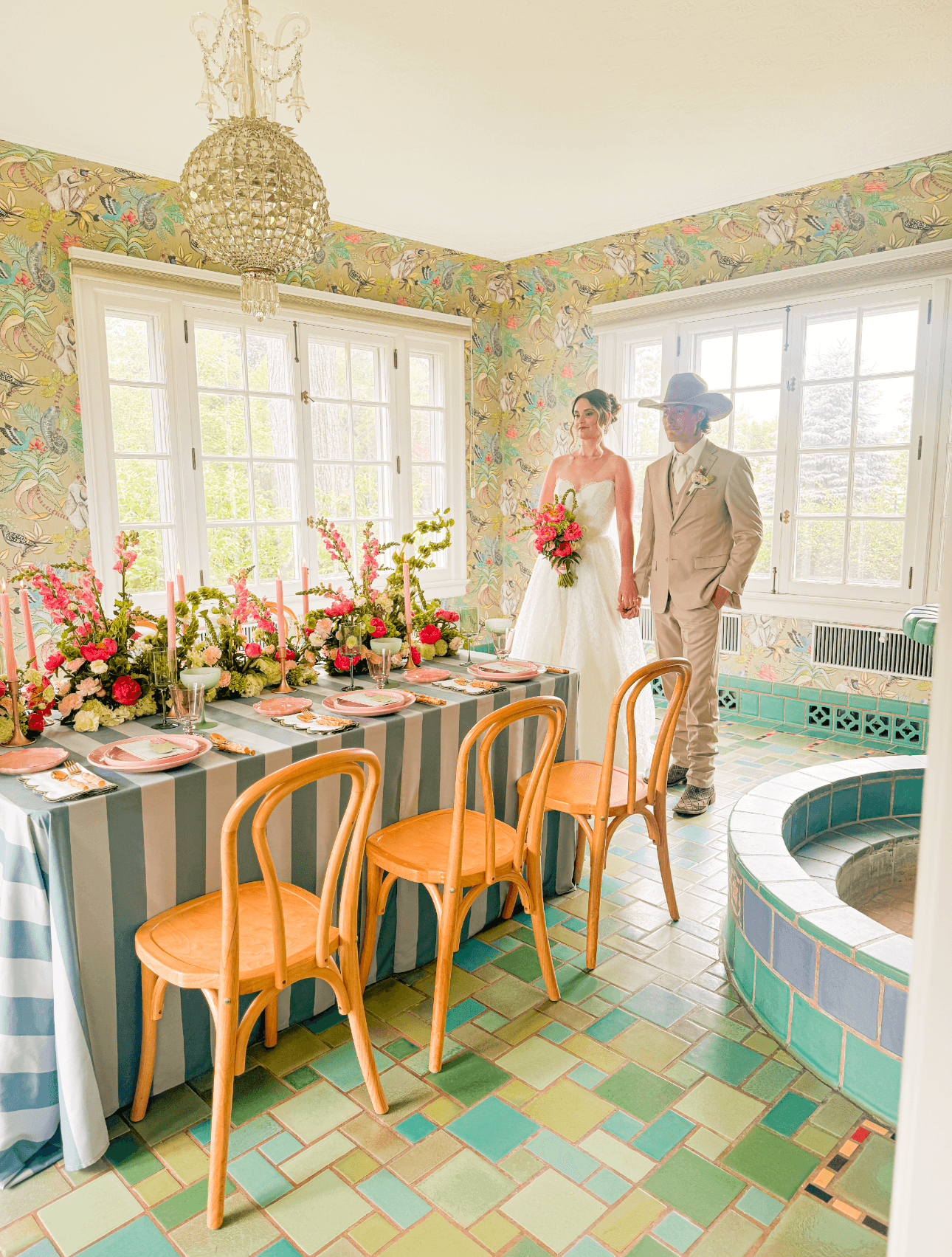 A bride and groom stand near a decorated table in a colorful room with large windows, floral wallpaper, and a small pool.