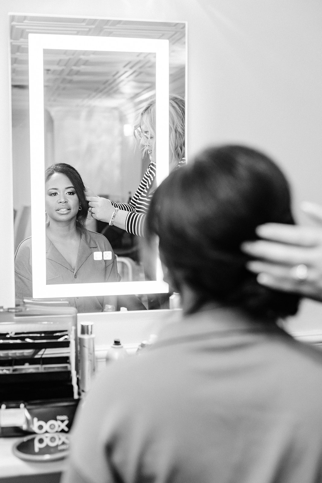 Person looking in mirror at woman getting hair styled, with hairdresser's hand visible, in black and white image.