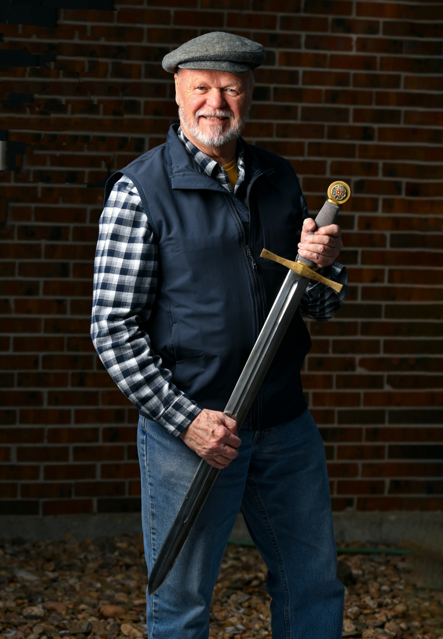 Older man with gray beard wearing a flat cap, plaid shirt, and vest, holding a large sword in front of a brick wall.