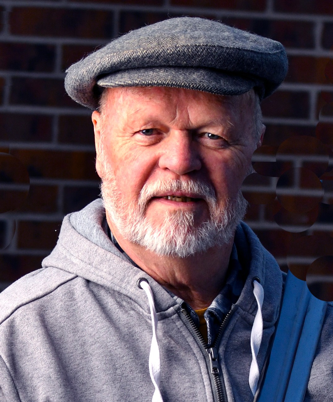Older man with a beard wearing a gray cap and hoodie, standing outdoors against a brick wall.