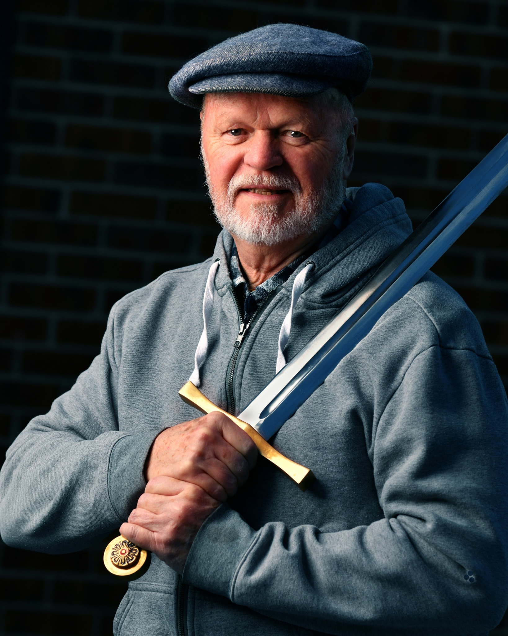 Older man with gray beard and mustache wearing a gray cap and hoodie, holding a sword with a gold hilt, standing outdoors.