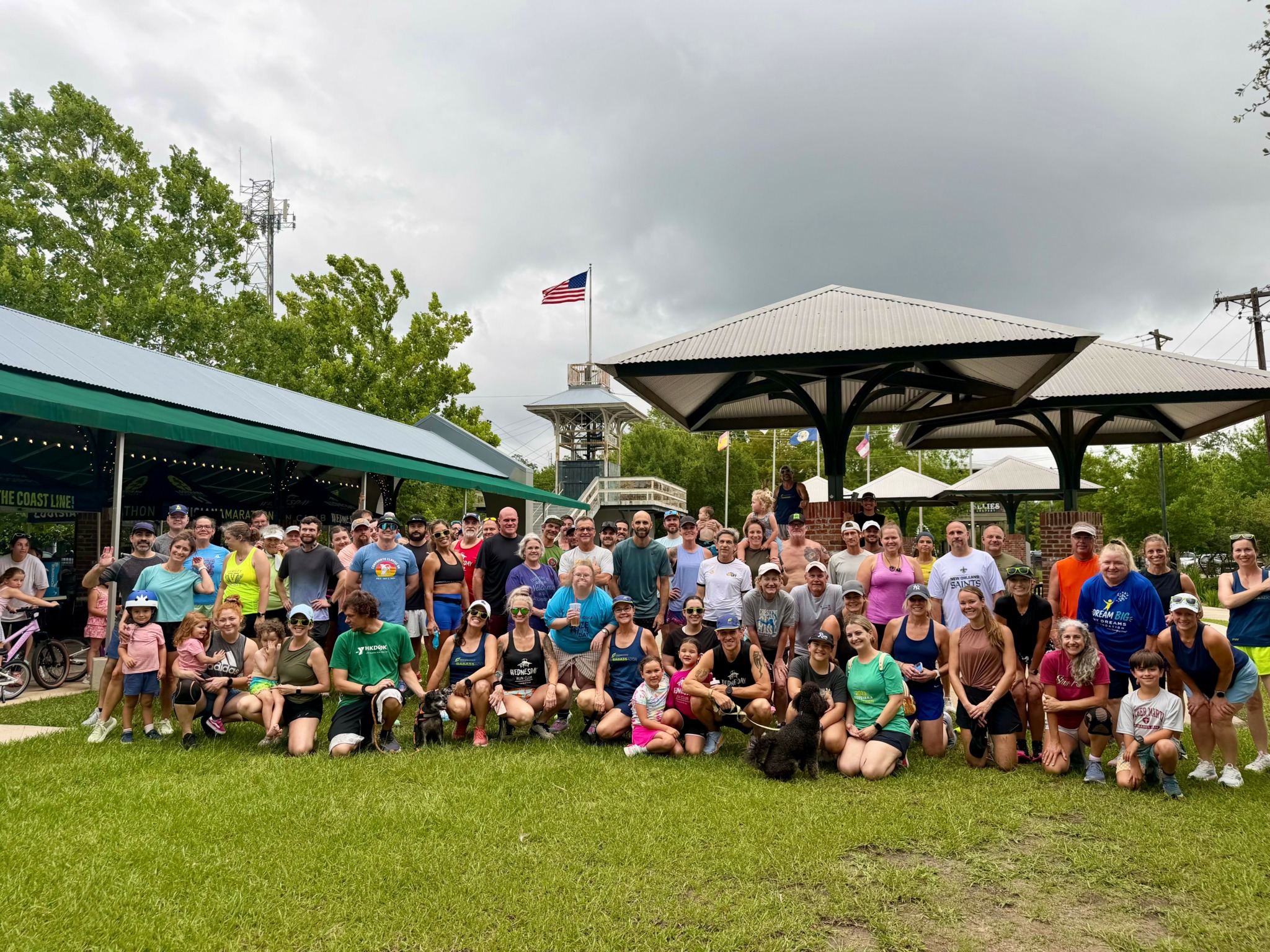 Large group of people, including children, gathered outdoors in a park with a pavilion and American flag, overcast sky.