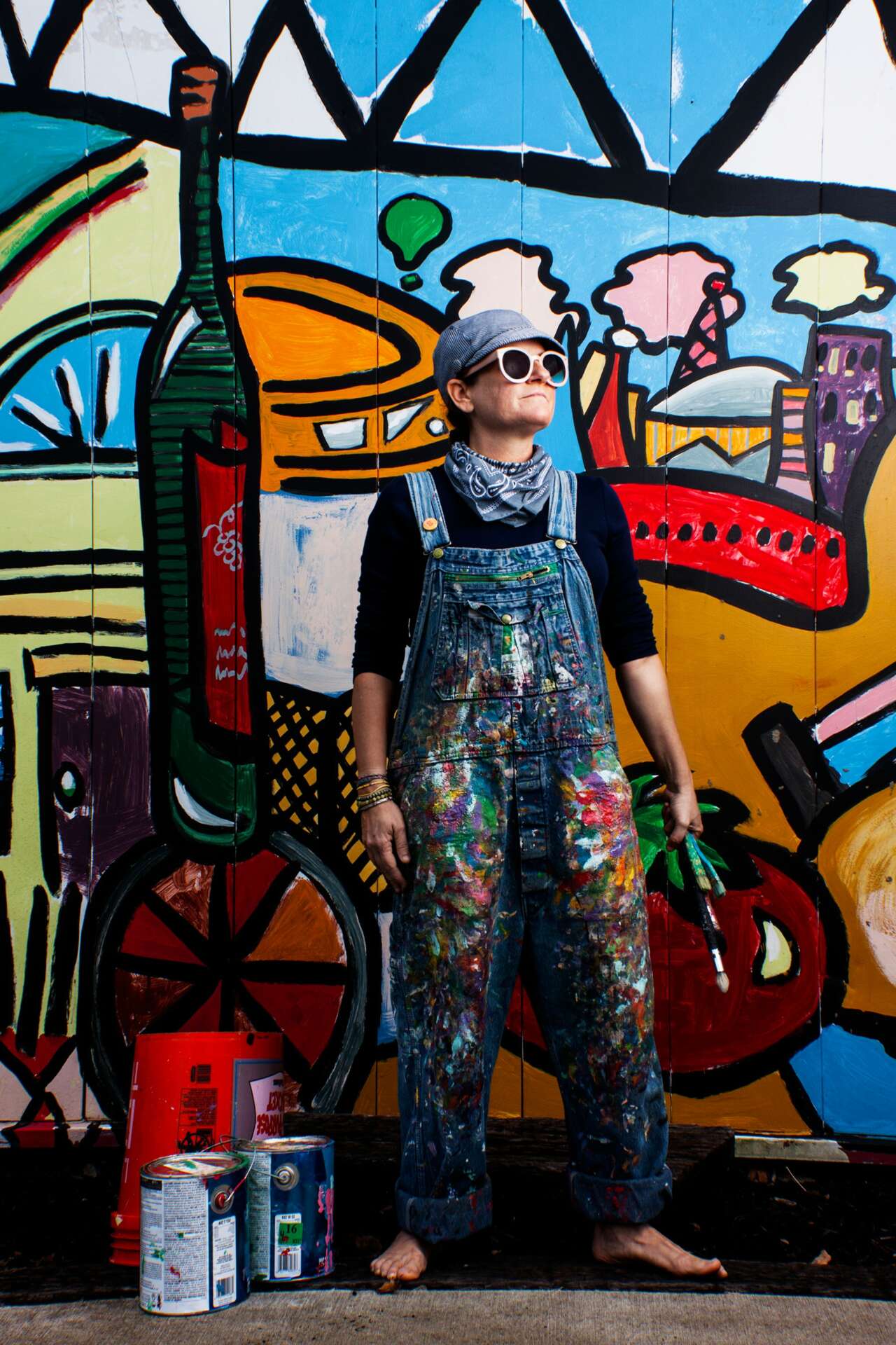 Person wearing sunglasses and overalls stands in front of a colorful graffiti mural, holding a spray paint can.