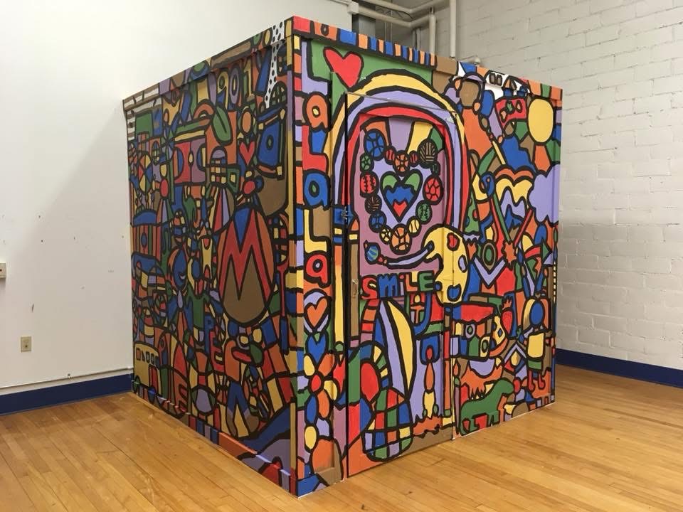 Colorful, abstract mural with various shapes, patterns, and a smiling face, covering a cube-shaped structure in a room.