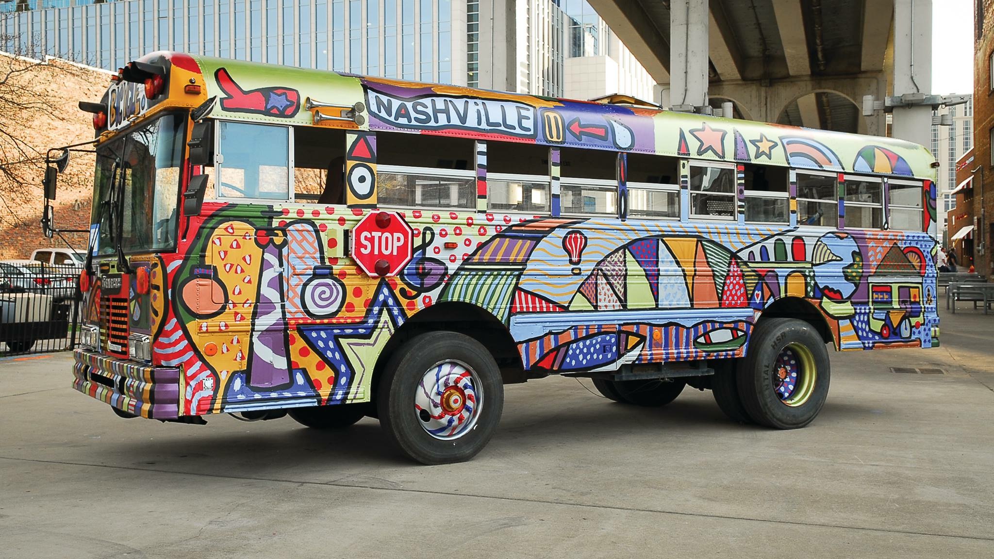 Colorful bus with abstract patterns, city signs, and decorative elements, parked under a bridge, with urban background.