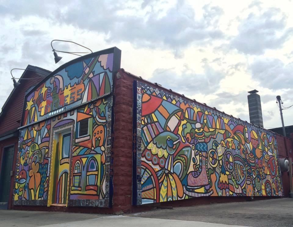 Colorful mural on building wall with abstract shapes, patterns, and figures, under cloudy sky, with a chimney and streetlamp nearby.