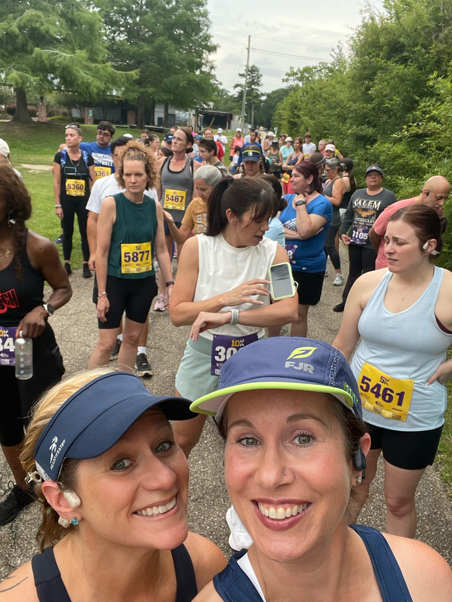 Two women smiling at the camera during a race event with many participants and trees in the background.