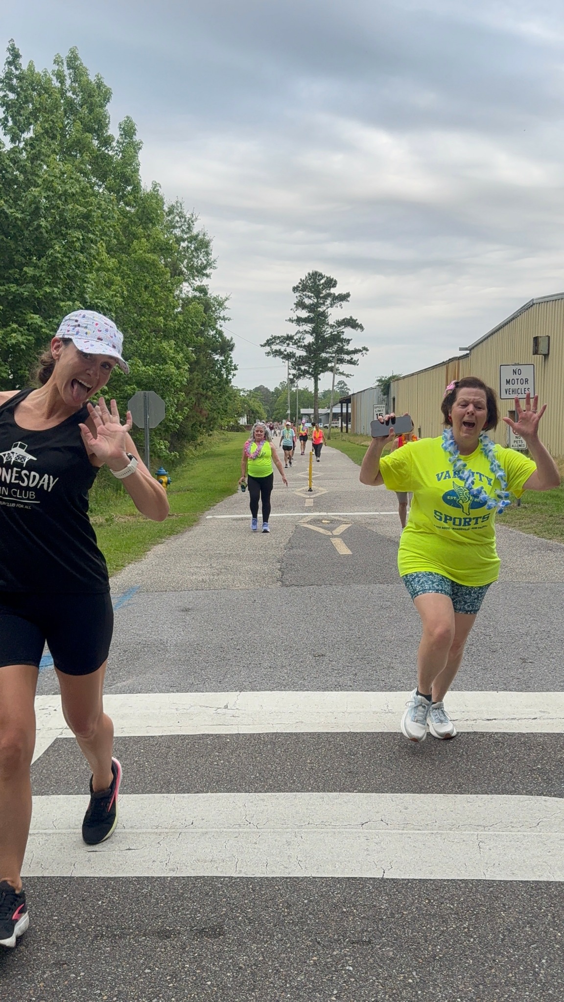 Two women smiling and waving at the camera while crossing a street during an outdoor event, with other people and trees in the background.