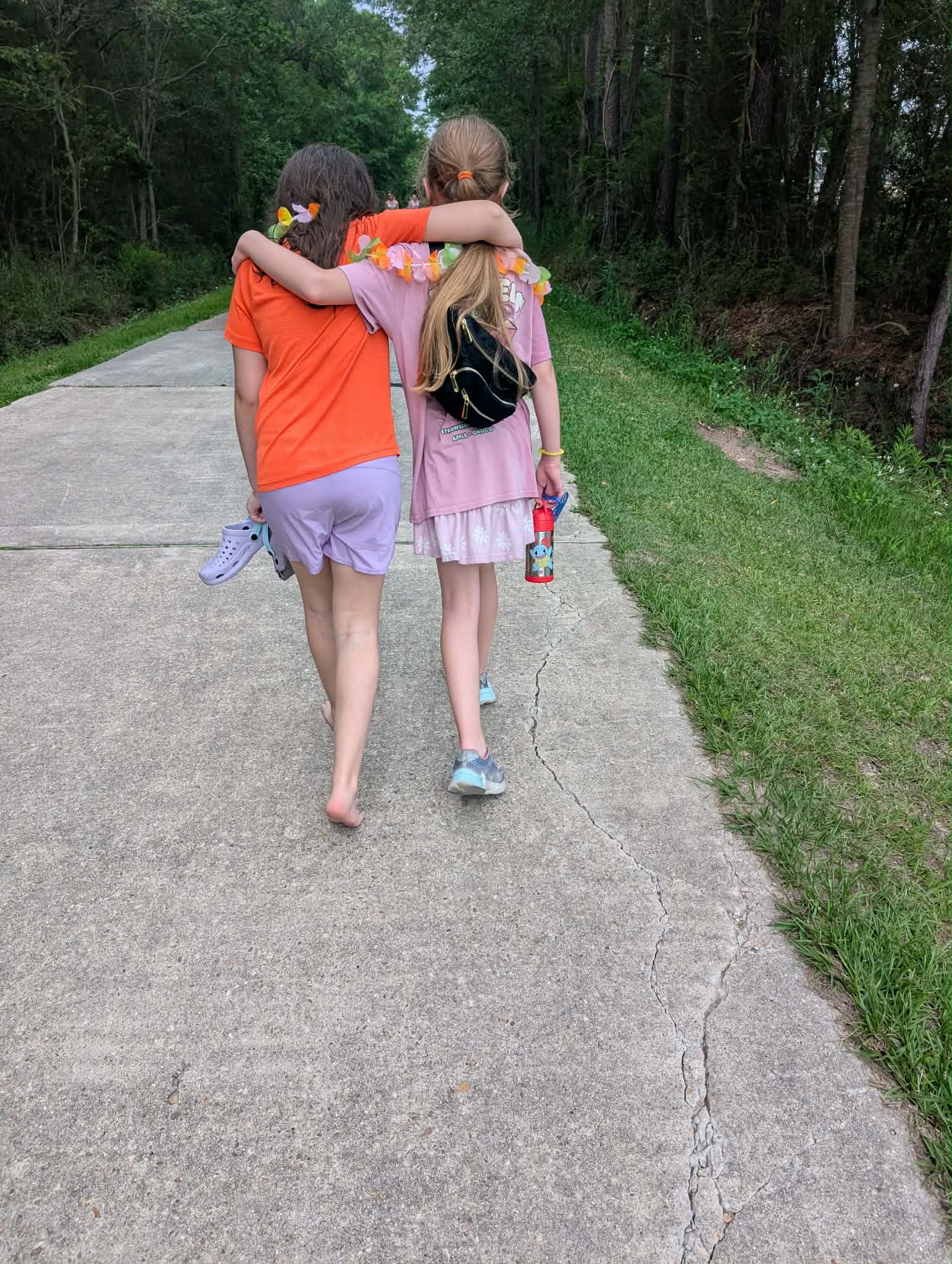 Two children walk arm-in-arm on a paved path in a forested area, one wearing an orange shirt and purple shorts, the other in a pink dress and backpack.