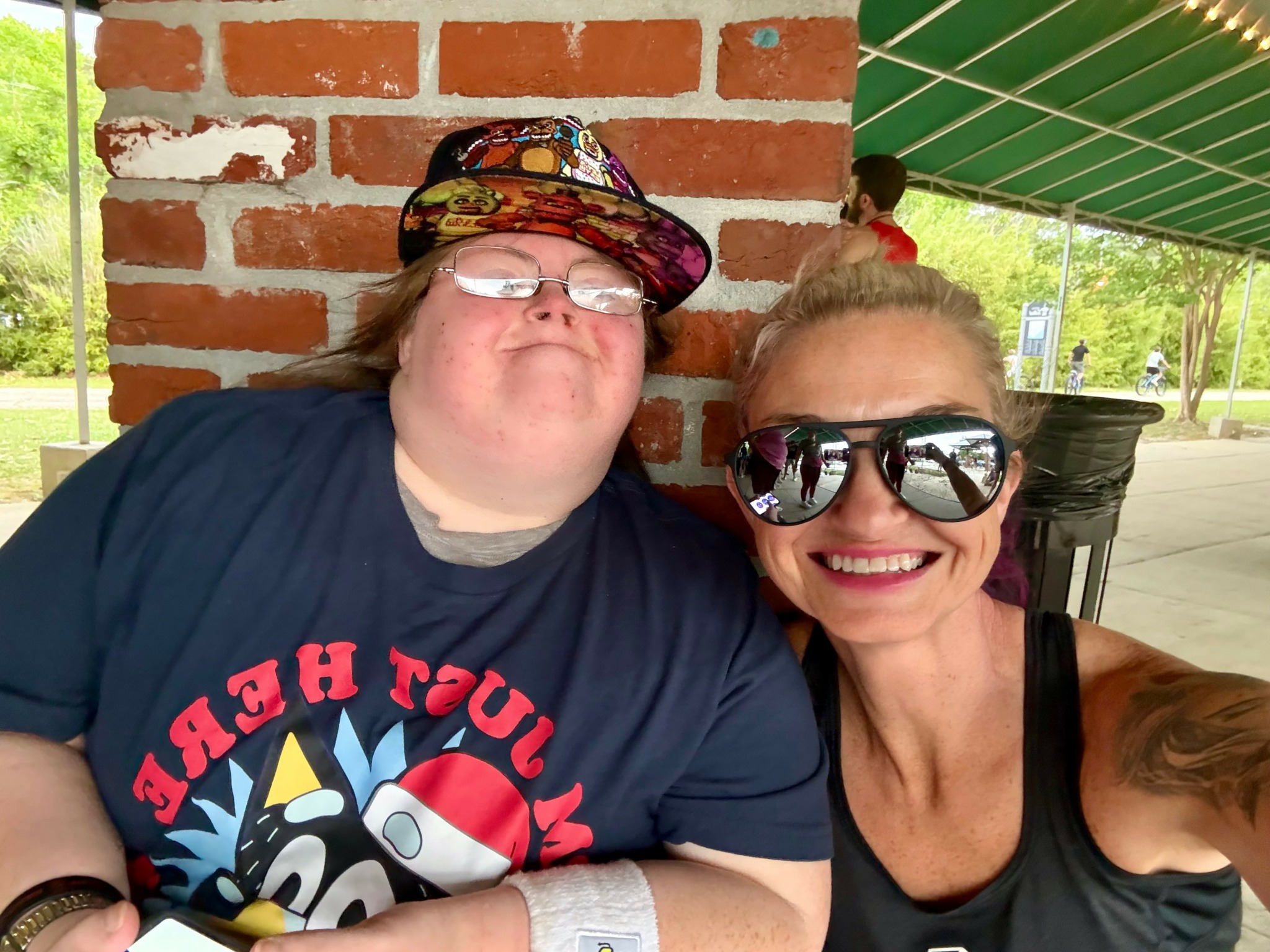 Two smiling women sitting outdoors, one wearing large sunglasses and a black top, the other wearing a colorful hat and glasses, with a brick wall behind them.