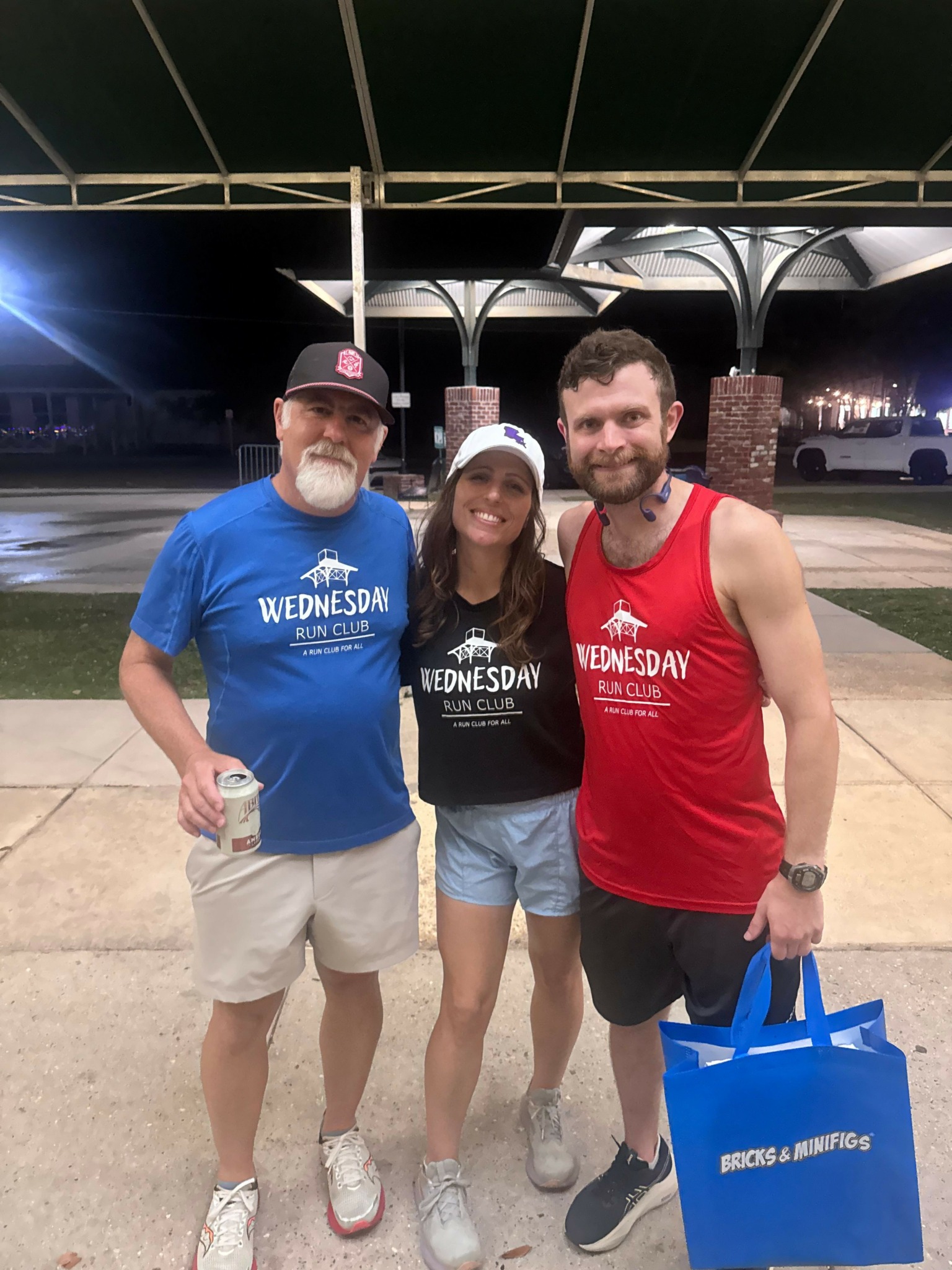 Three people standing outdoors at night, one holding a blue bag, all smiling, with a street and parked cars in background.