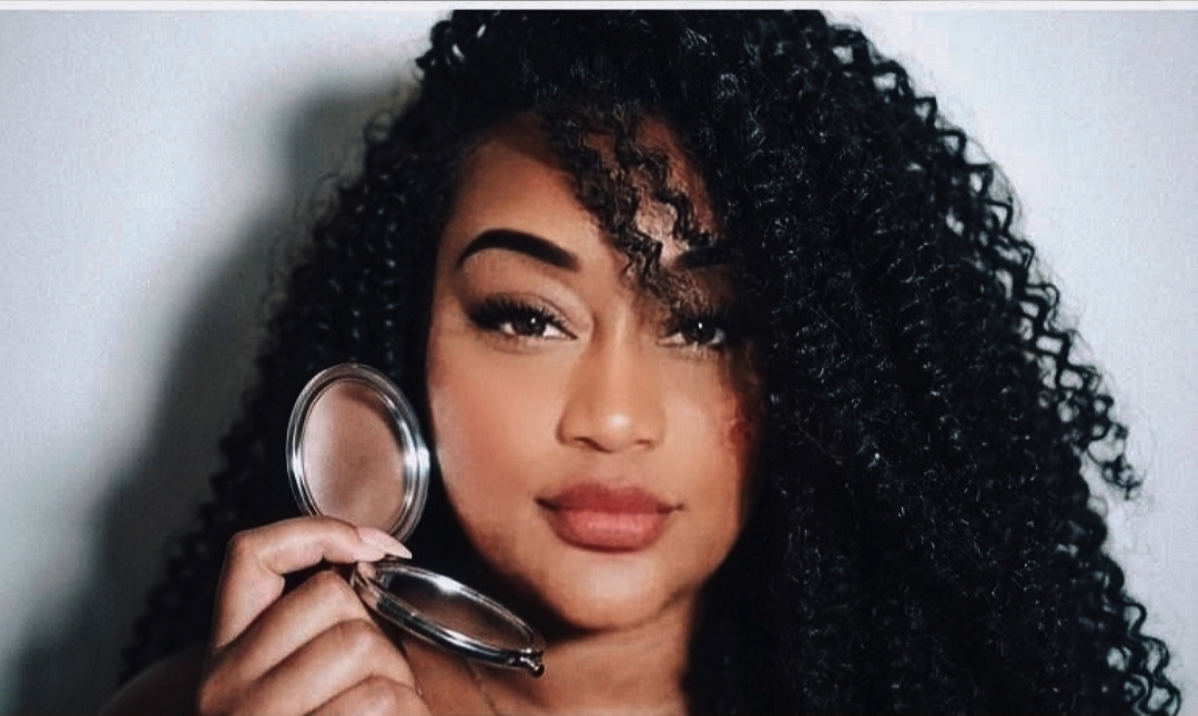 Woman with curly hair holding a mirror close to her face, looking at the camera.