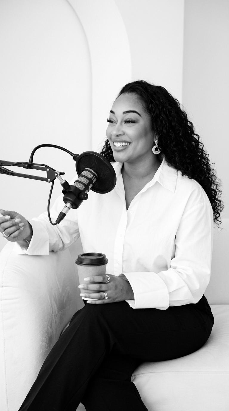 Woman with curly hair speaking into a microphone, holding a coffee cup, smiling, in a white shirt, black pants, in a studio.