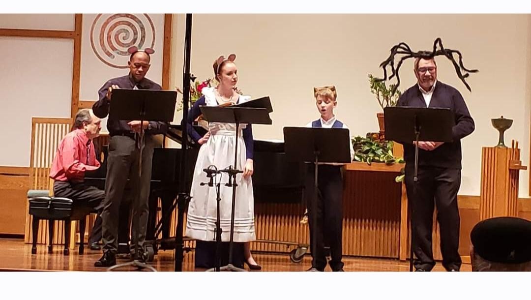 Five people stand on stage with music stands, performing in a room with wooden decor and a plant.