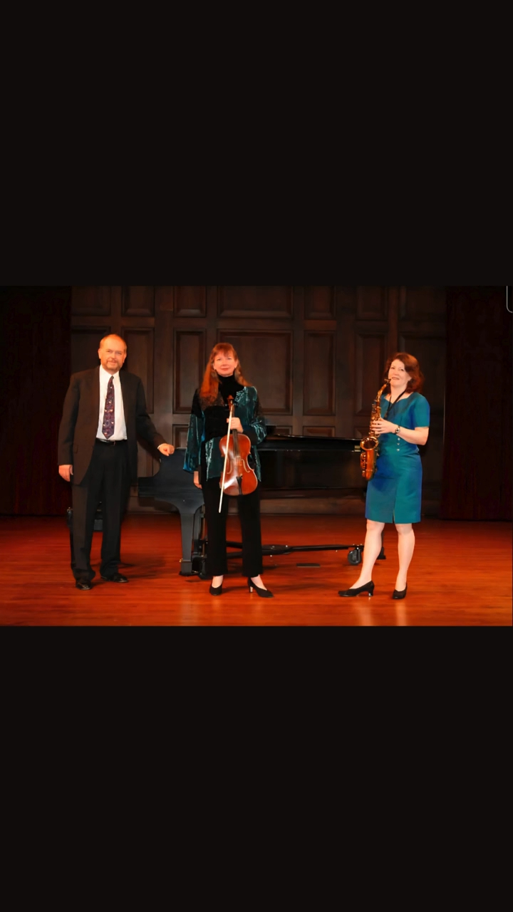 Three people standing on a stage with wooden paneling, two women and one man, engaged in conversation.