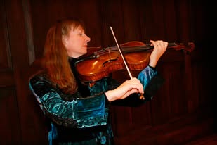 Woman playing violin with a bow, wearing a blue velvet dress, against a dark wooden background.