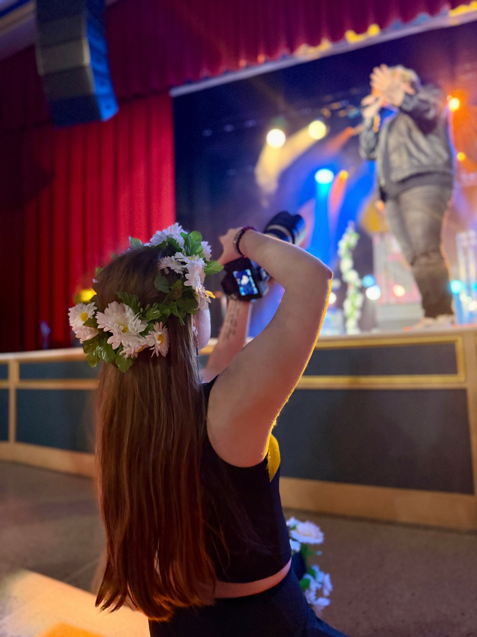 Person with long hair and a flower crown taking a photo of a performer on stage with a microphone, colorful lights in background.