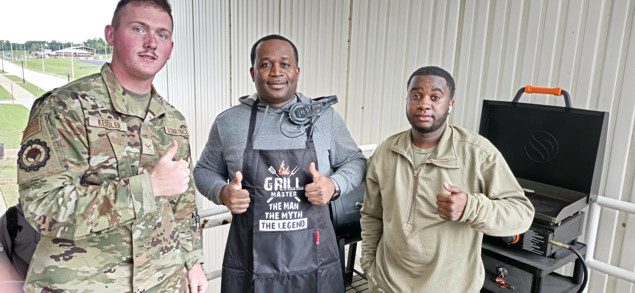 Three men standing indoors, one in military uniform, one in apron, and one in casual clothing, with a computer in the background.