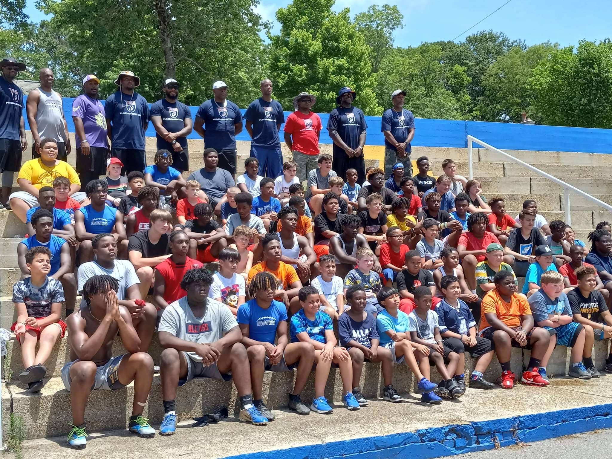 Group of children and adults sitting and standing on stadium steps outdoors, with trees in background.