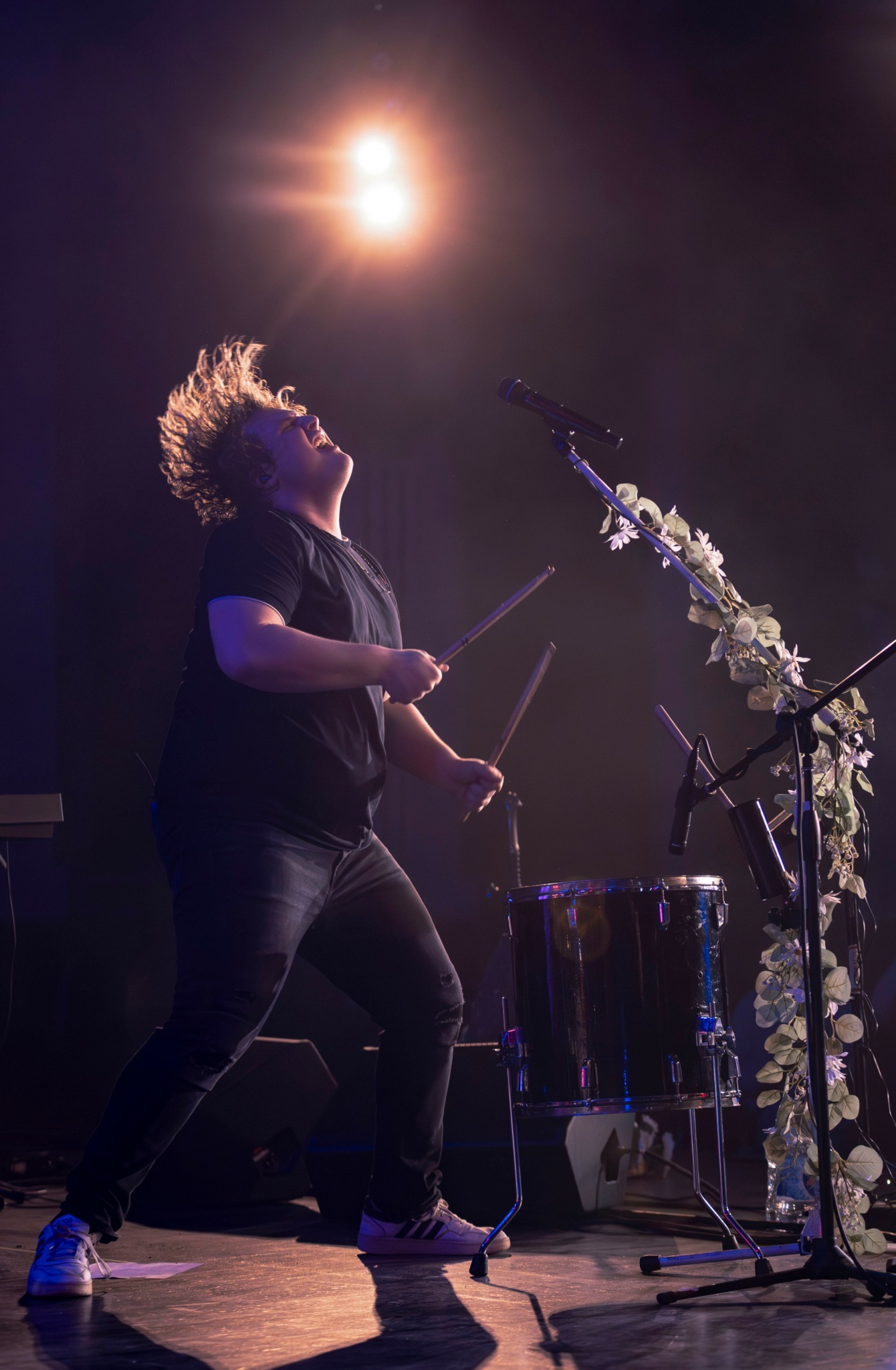 Performer with curly hair playing drums on stage under bright light, with microphone and floral decoration nearby.