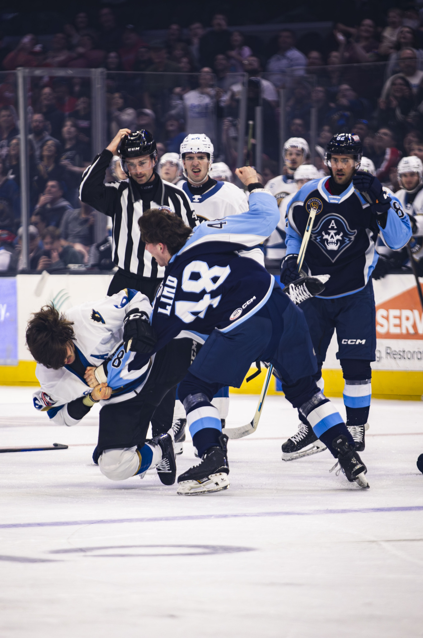 Two hockey players fight on ice, surrounded by referees and other players, with spectators in the background.