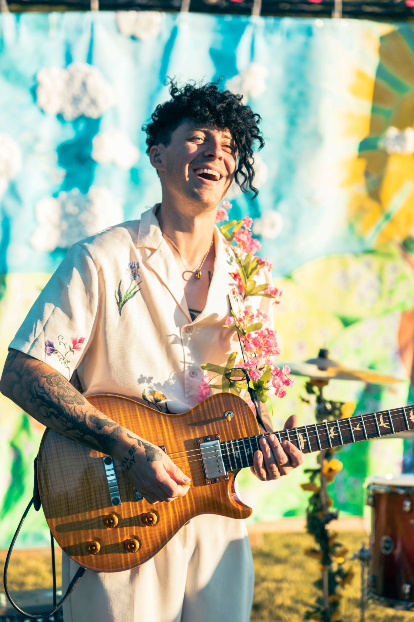 Person with curly hair playing an electric guitar outdoors, smiling, with colorful background and flowers.