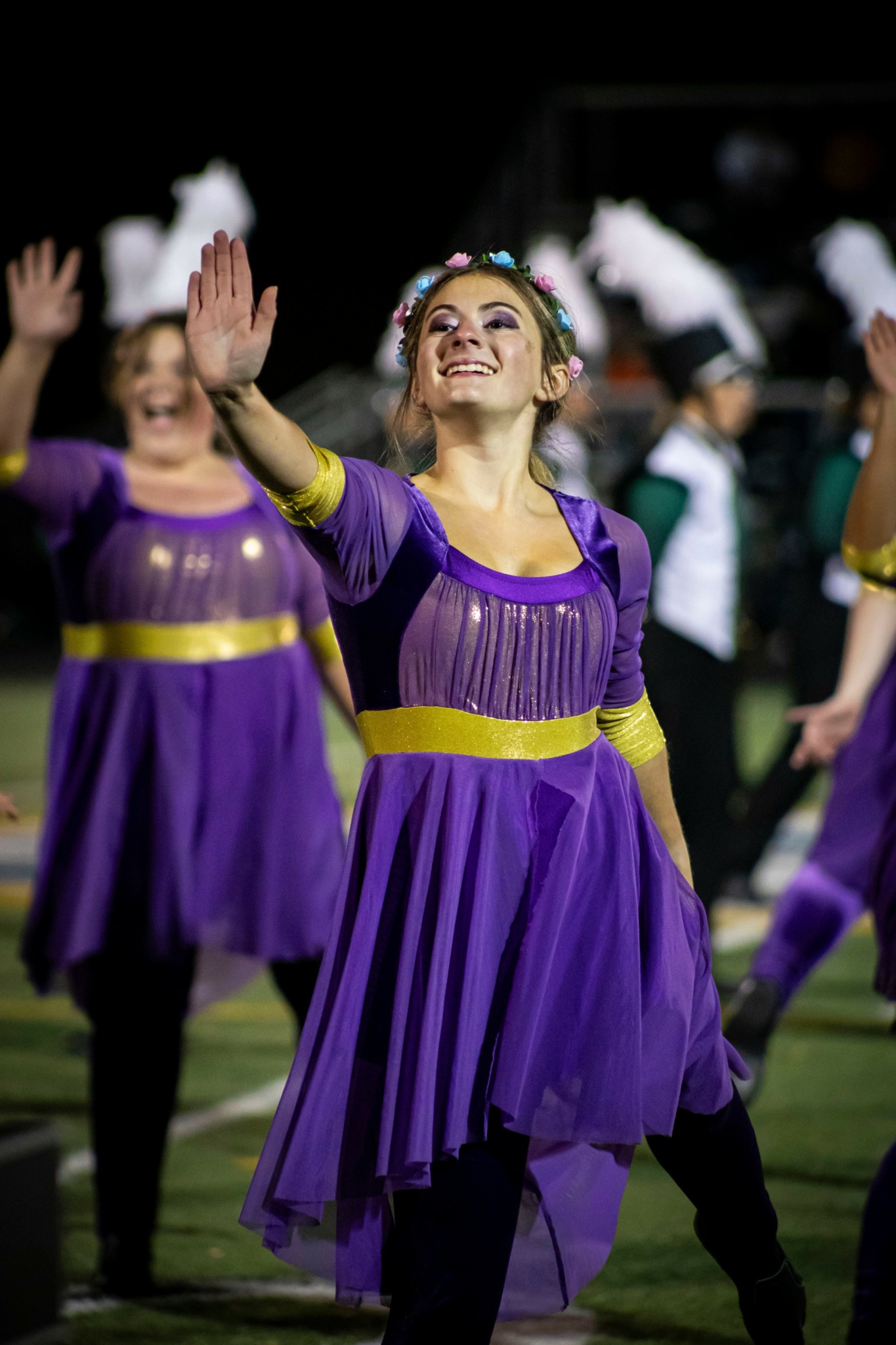 A smiling woman in a purple dress with yellow accents raises her hand during a performance, surrounded by others in similar costumes.