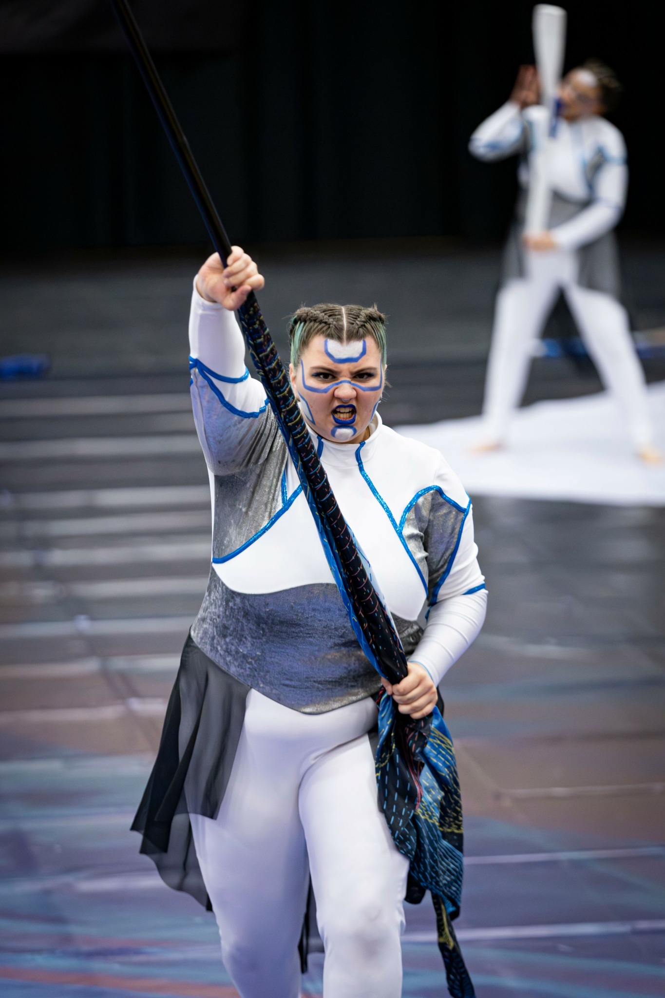 Performer in costume holding a staff, with another performer in background on stage, dark backdrop, and steps.
