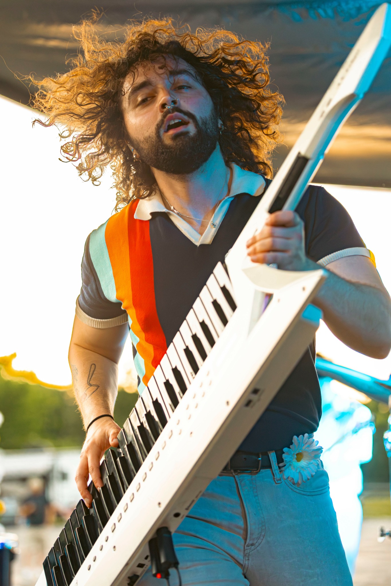 Person with curly hair playing a white keyboard outdoors, wearing a dark shirt with rainbow stripes, holding a tablet.