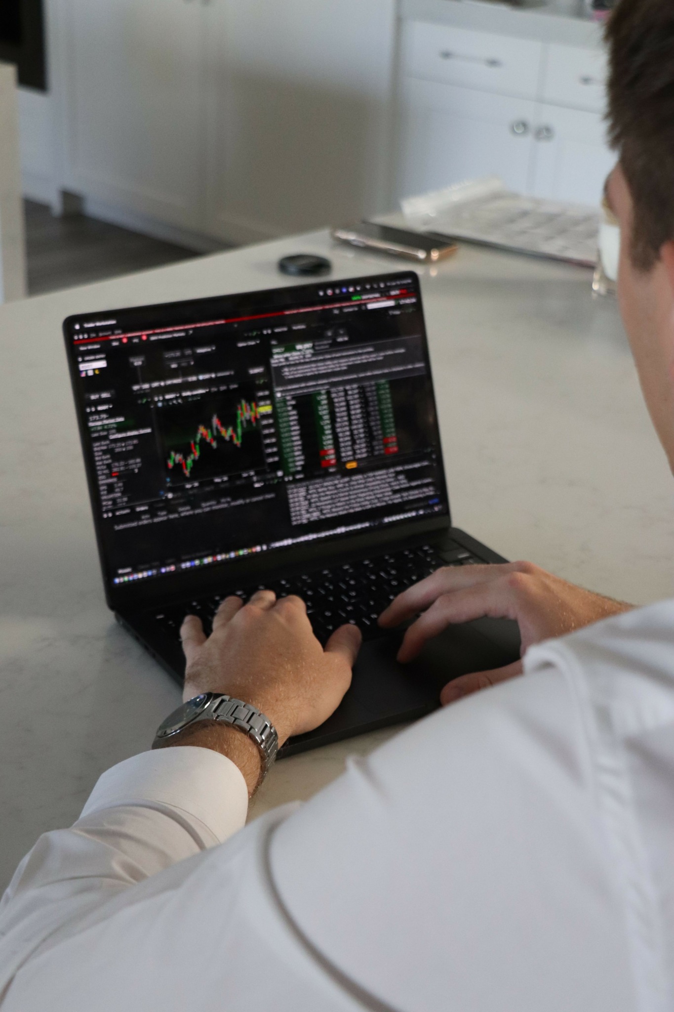 Person using a laptop with stock trading charts and data displayed on the screen, sitting at a table in a bright room.
