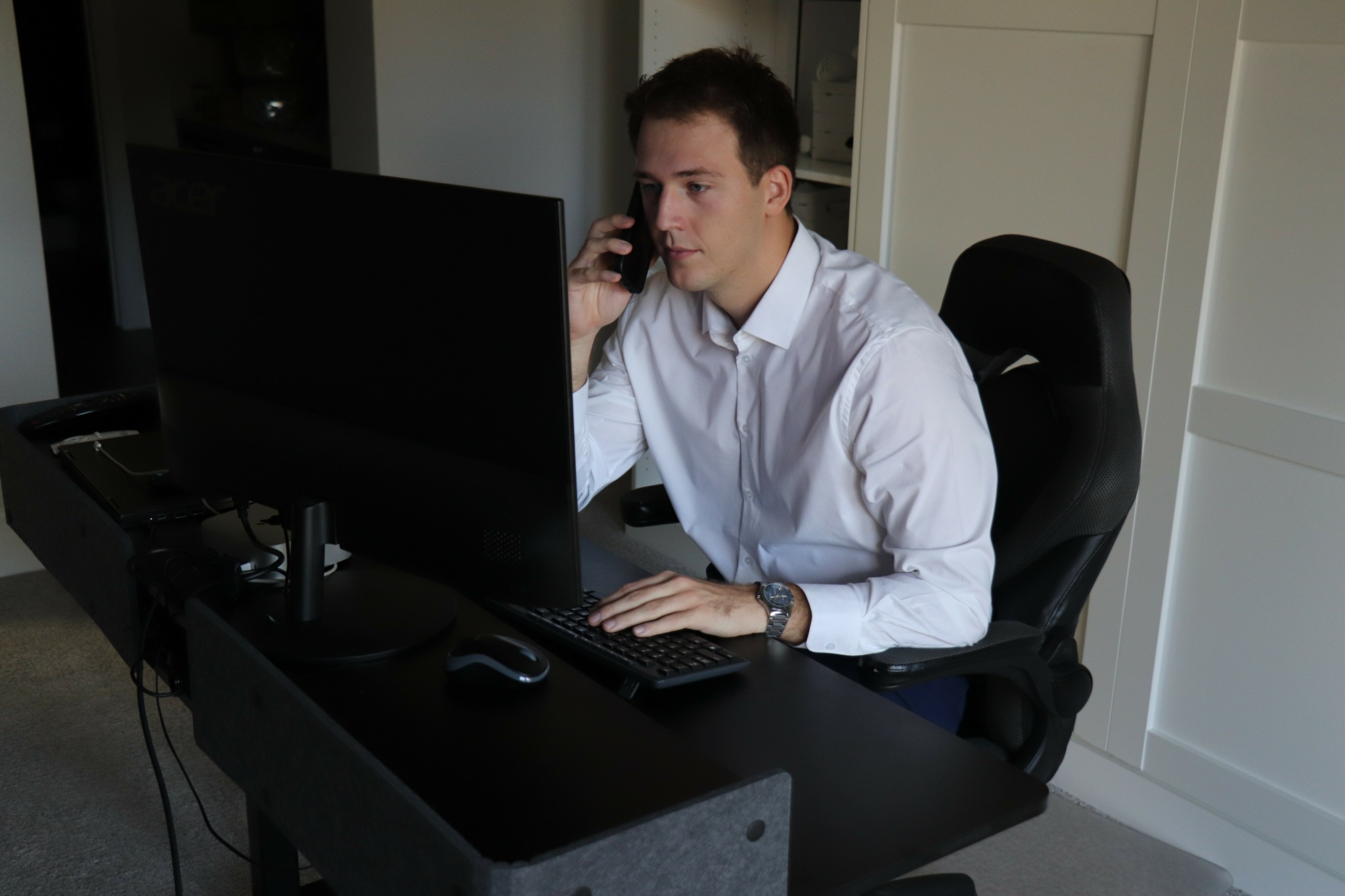 Man in white shirt sitting at desk, talking on phone, with computer monitor, keyboard, and mouse, in a room with white cabinets