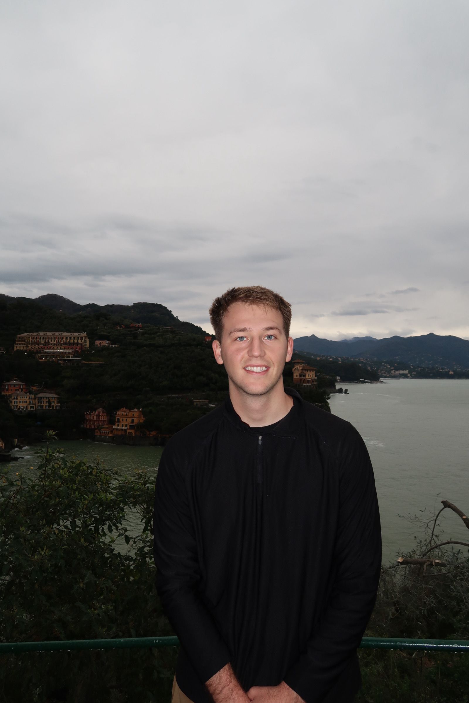 Person standing outdoors near water with trees and cloudy sky in background.