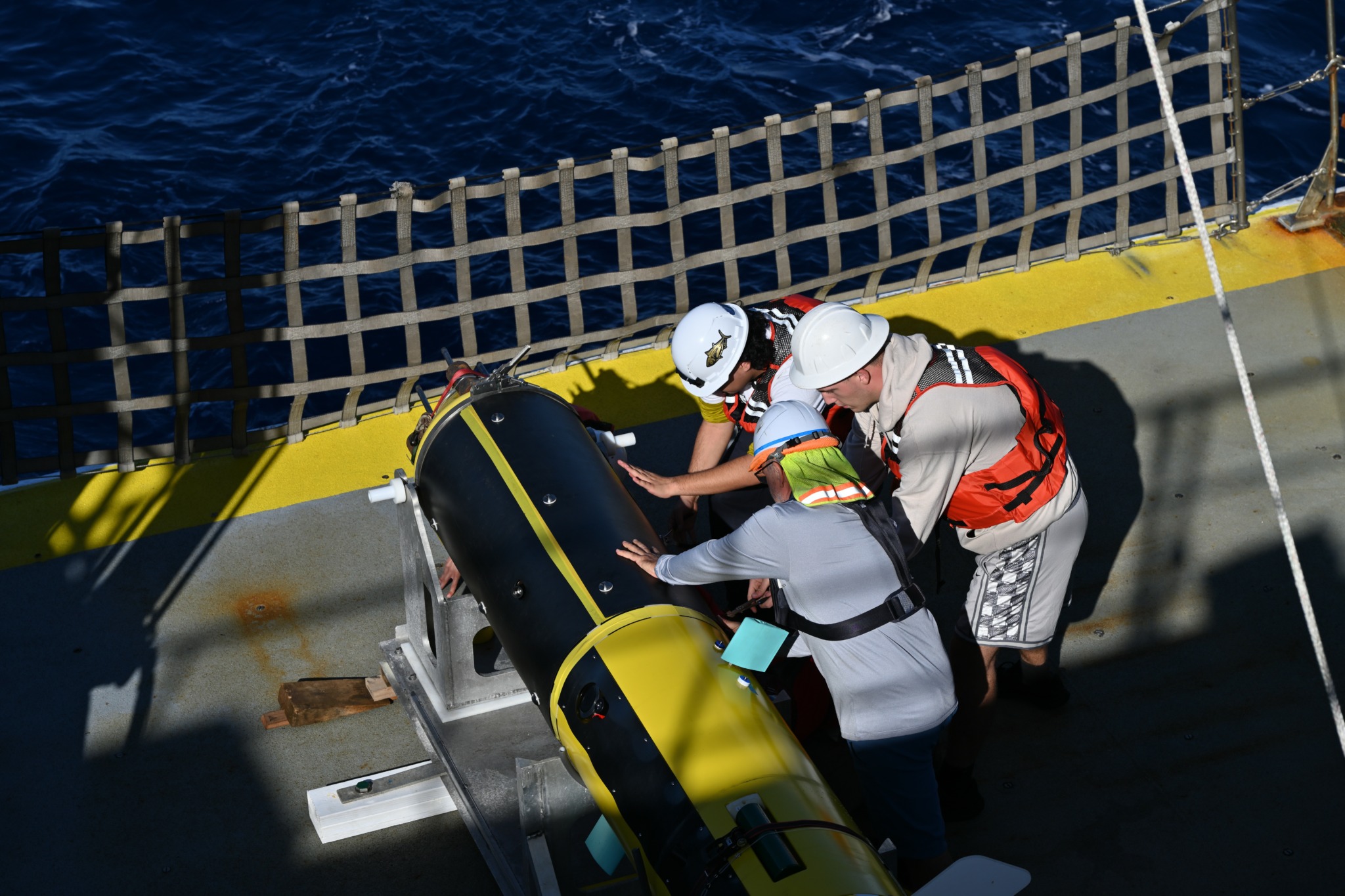 Three people in safety gear work together on a yellow boat with a black and yellow device, near a safety net, on water.