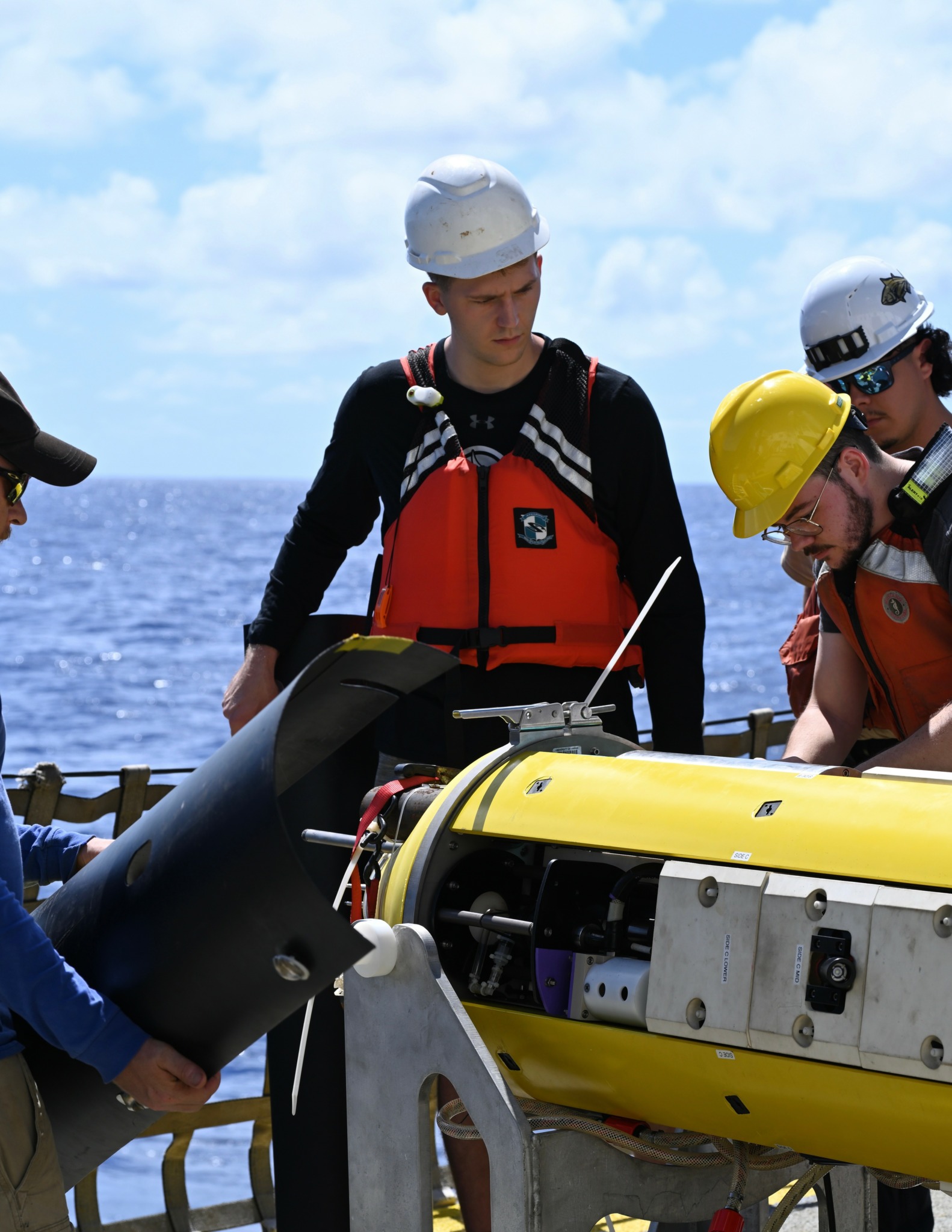 Group of people in safety gear working on a yellow underwater robot on a boat, with ocean and sky in background.