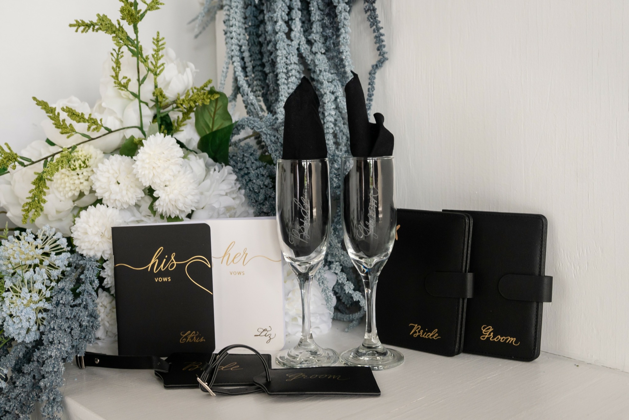 Two empty champagne flutes in front of a floral arrangement and black boxes with gold writing, on a white surface.