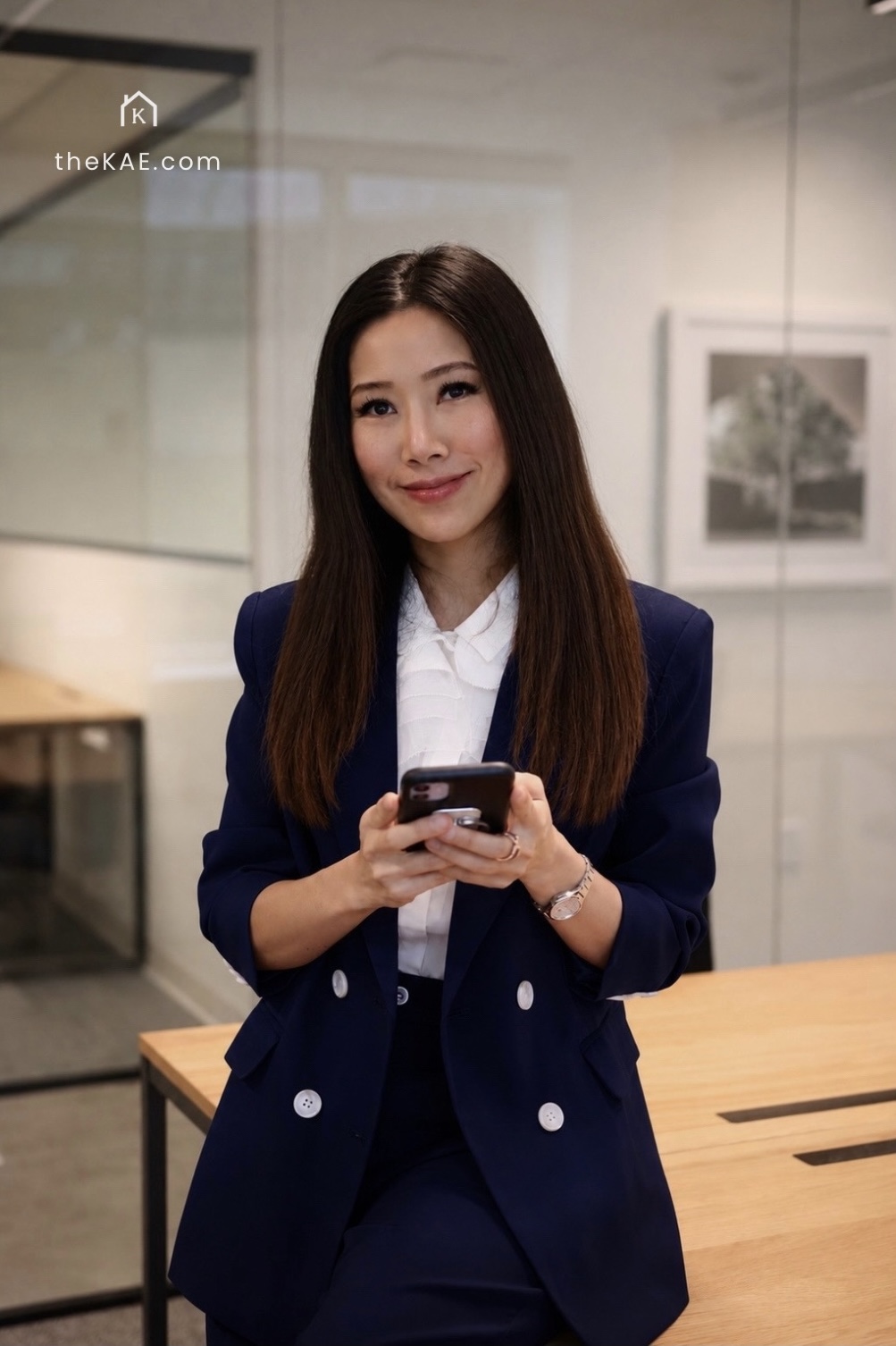 Woman in navy suit holding a smartphone, standing in an office with framed pictures on the wall.