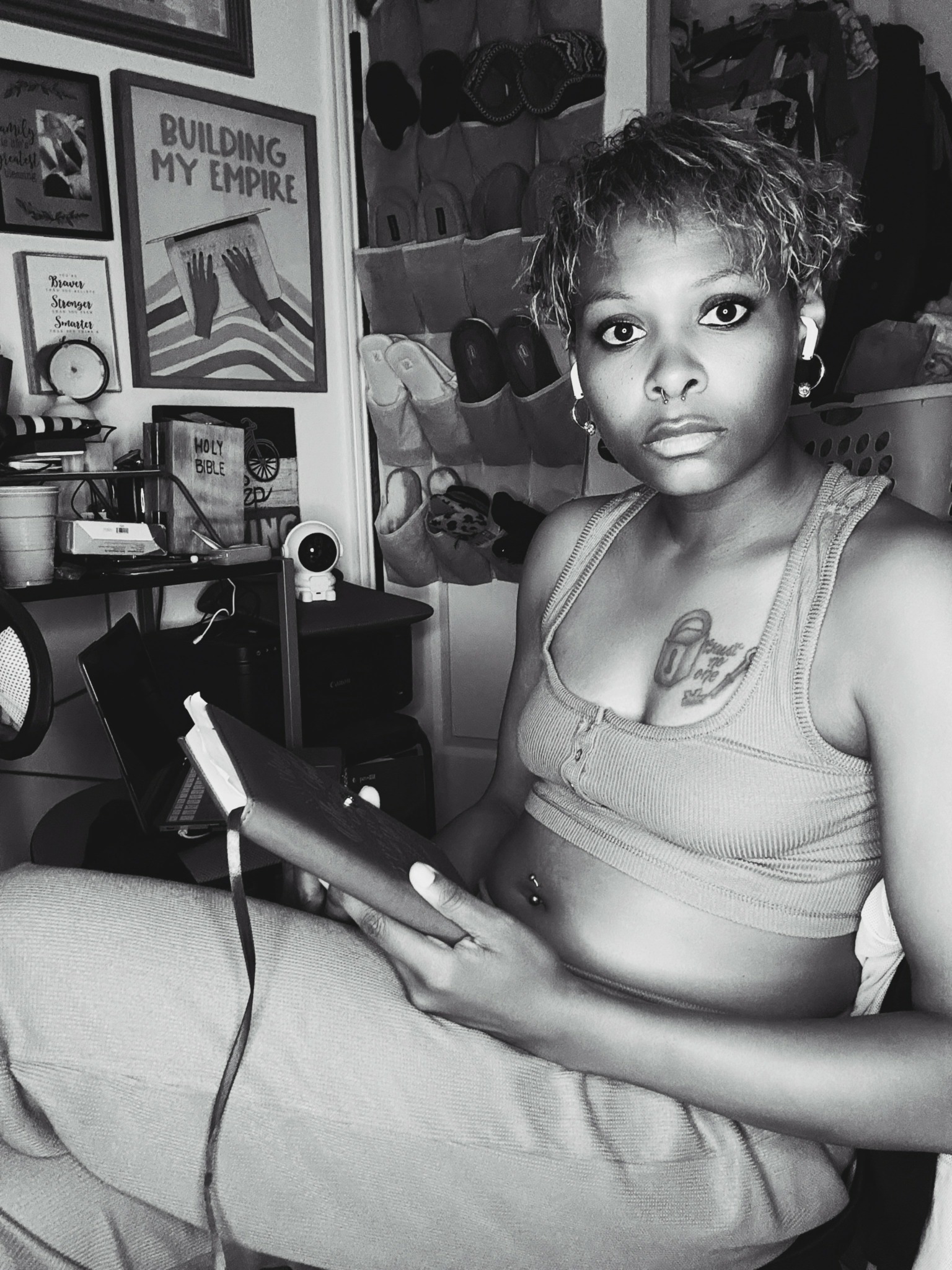 Young woman with short curly hair holding a tablet, sitting in a room with shelves and framed pictures behind her.