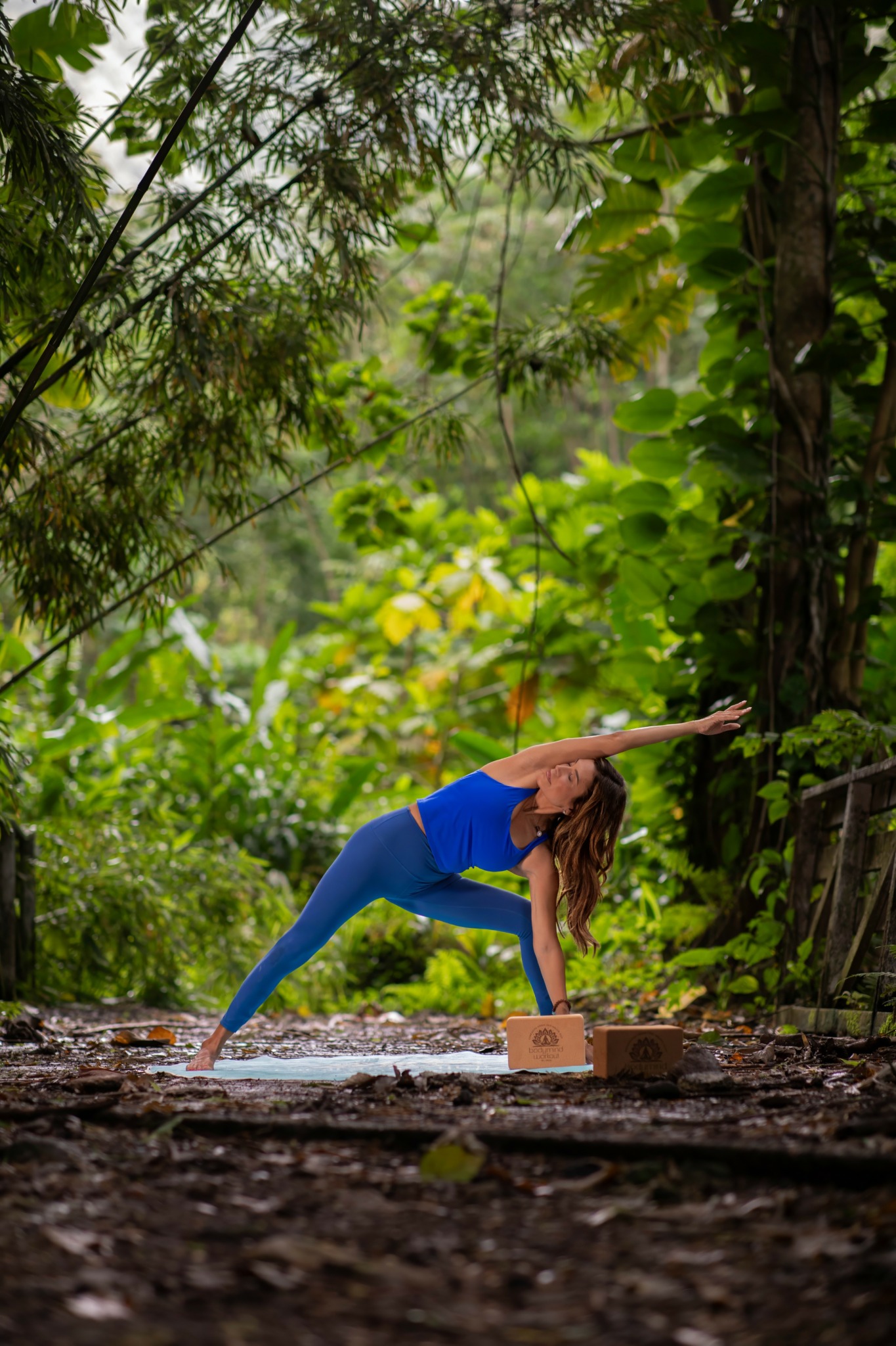Person practicing yoga outdoors in a forest, stretching with one arm extended and one leg bent, on a yoga block.